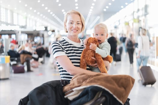 Mother traveling with child, holding his infant baby boy at airport terminal waiting to board a plane. Travel with kids concept