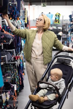 Casualy dressed mother choosing sporty shoes and clothes products in sports department of supermarket store with her infant baby boy child in stroller