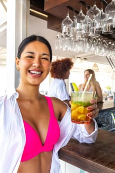 Vertical portrait of happy young latina woman showing mojito cocktail looking at camera at a beach bar in summertime. Vacation concept.