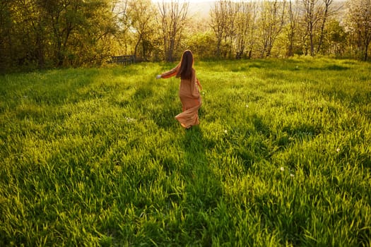 an attractive, slender, red-haired woman stands far away in a wide, green field during sunset in a long orange dress enjoying unity with nature and relaxation standing with her back to the camera. High quality photo