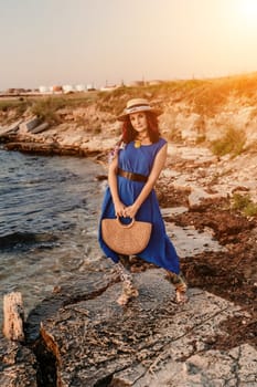 A woman in a dress, hat and with a straw bag is standing on the beach enjoying the sea. Happy summer holidays.