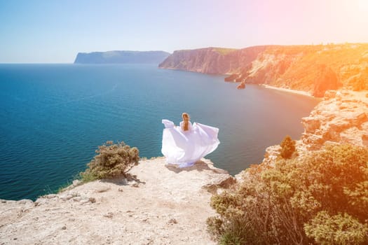 woman sea white dress. Blonde with long hair on a sunny seashore in a white flowing dress, rear view, silk fabric waving in the wind. Against the backdrop of the blue sky and mountains on the seashore