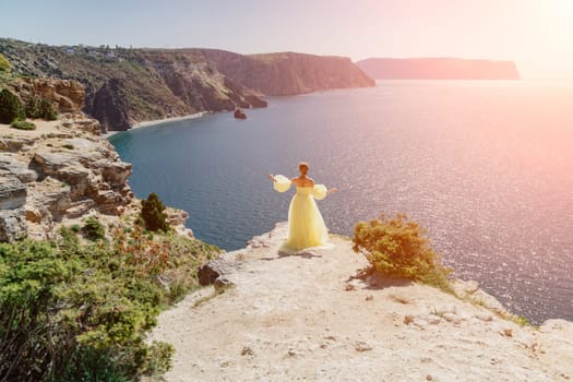 Woman yellow dress sea. Side view Young beautiful sensual woman in yellow long dress posing on a rock high above the sea at sunset. Girl in nature against the blue sky.