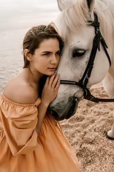 A woman in a dress stands next to a white horse on a beach, with the blue sky and sea in the background