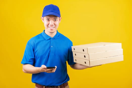 Excited delivery service man smiling wearing blue uniform hold paper containers for takeaway pizza boxes packet and show smartphone blank screen, studio shot isolated on yellow background