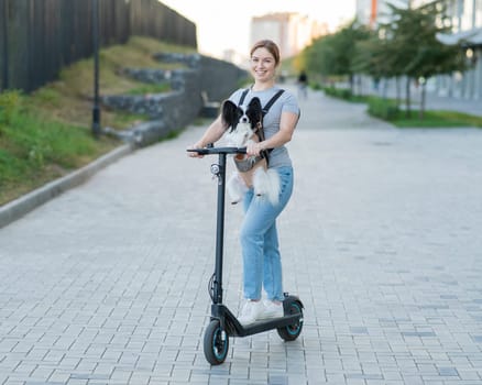 A woman rides an electric scooter with a dog in a backpack. Pappilion Spaniel Continental in a sling