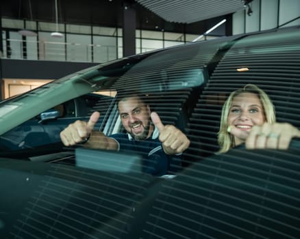 Happy caucasian couple is sitting in a new car in a car dealership. Man showing thumbs up