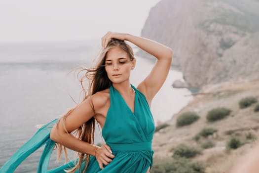 Side view a Young beautiful sensual woman in a mint long dress posing on a volcanic rock high above the sea during sunset. Girl on the nature on overcast sky background. Fashion photo