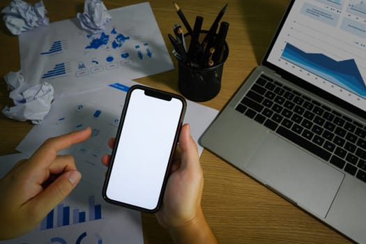 Hand holding smartphone, sitting in front of laptop at messy office desk. Close up view, blank screen for advertising text message.