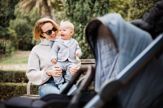 Mother sitting on bench in urban park, laughing cheerfully, holding her smiling infant baby boy child in her lap having baby stroller parked by their site