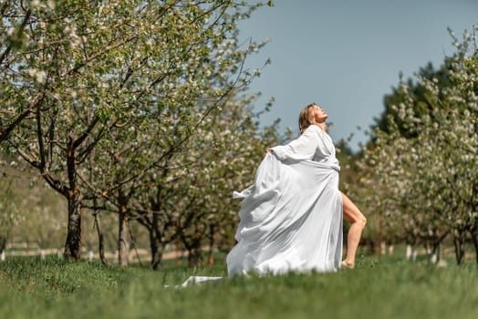 Blond blooming garden. A woman in a white dress walks through a blossoming cherry orchard. Long dress flies to the sides