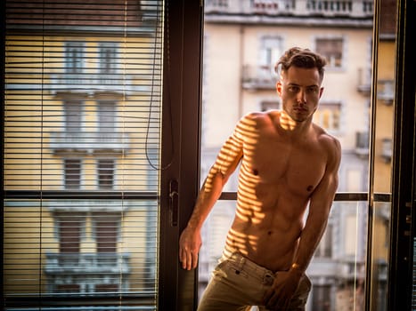Portrait of sexy shirtless man covered with shadow stripes from window, looking at camera