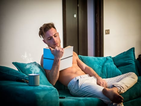 Handsome shirtless athletic young man reading book while sitting on couch