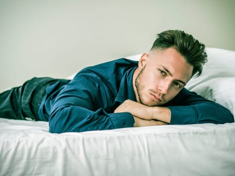 Sexy male model laying alone on bed in his bedroom, looking at camera with a seductive attitude,, wearing blue elegant shirt