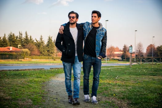 Two handsome casual trendy young men, 2 friends, in an urban park walking and chatting together