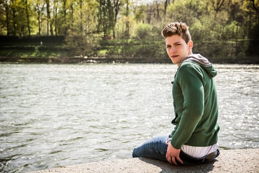 Three-quarter length of contemplative light brown haired young man wearing green hooded-shirt and denim jeans sitting on wall beside picturesque river in Turin, Italy