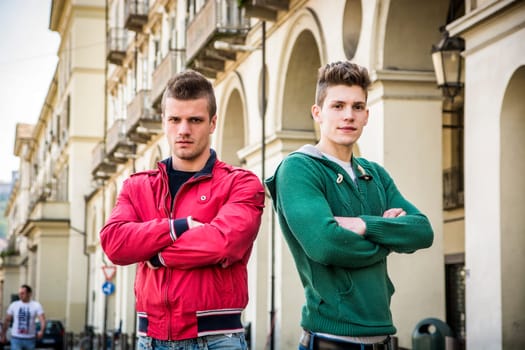 Two young male friends standing in European city center, looking at camera with arms crossed on chest