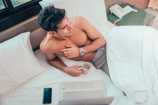 Naked young man with muscular body on bed with mug or cup in hand with coffee or tea, looking at laptop computer screen