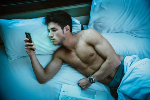Handsome young man in bed typing on cell phone, sending text message or dialing number or reading web pages, late at night