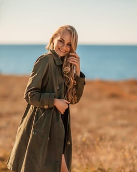 Portrait blonde sea cape. A calm young blonde in an unbuttoned khaki raincoat stands on the seashore, under the raincoat there is a black skirt and top.