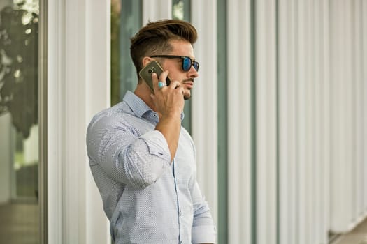 Handsome man calling on cellphone, wearing elegant shirt posing outside on urban background.