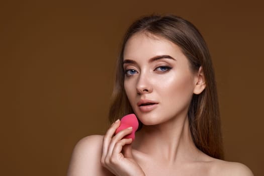 smiling young woman applying foundation on skin with beauty blender isolated on beige background