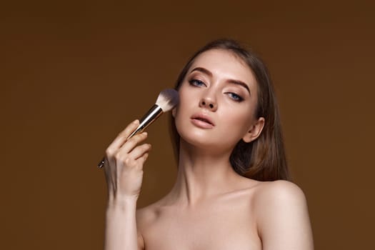 young woman applying blush on cheeks with make-up brush on beige background. copy space