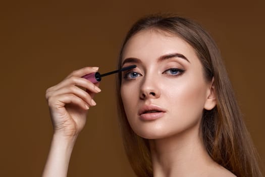 Beautiful woman applying black mascara on long thick eyelashes with brush on beige background