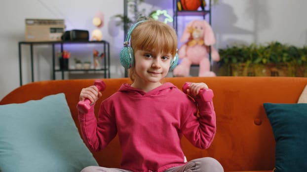 Young child kid girl lifting dumbbells while doing biceps exercises, enjoying training. Female teen toddler wearing wireless headphones listening music at home in room sits on sofa. Sport and fitness