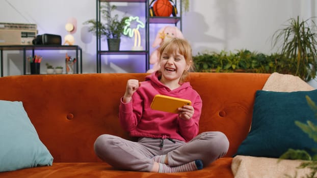 Worried funny child kid girl enthusiastically playing online racing or shooter video games on smartphone. Female teen toddler using smartphone gadget app with drive simulator at home sits on sofa