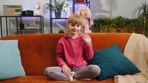 Young blonde child kid girl smiling friendly at camera and waving hands gesturing hello, hi, greeting or goodbye, welcoming with hospitable expression. Teen toddler at home in living room sits on sofa