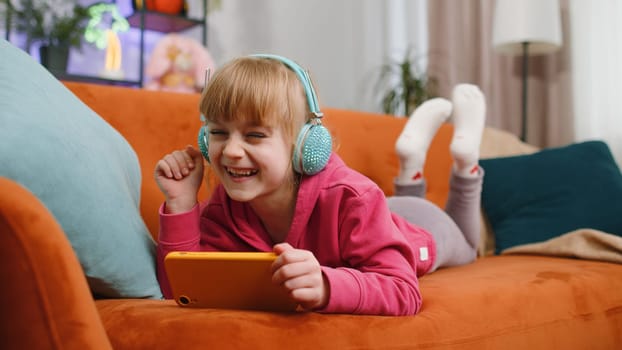 Worried funny child kid girl enthusiastically playing online racing or shooter video games on smartphone. Female teen toddler using smartphone gadget app with drive simulator at home lying on sofa