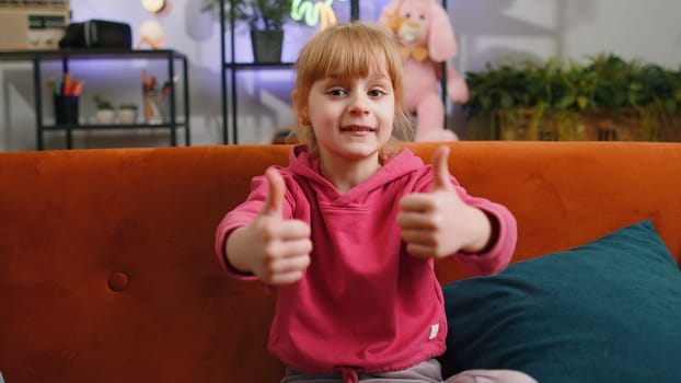 Like. Happy young blonde child kid girl looking approvingly at camera showing thumbs up, like sign positive something good, positive feedback. Female teen toddler at home in living room sits on sofa