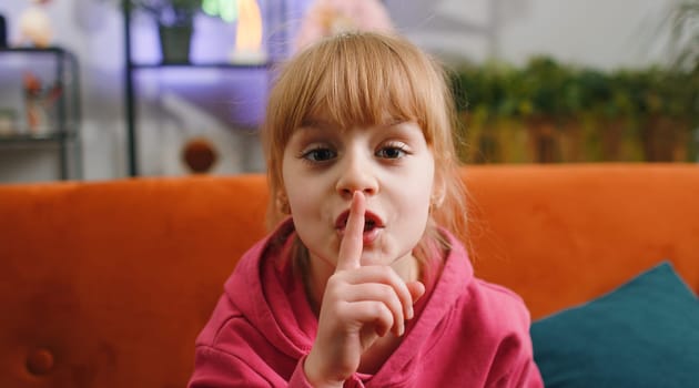 Shh be quiet please. Young blonde child kid girl presses index finger to lips makes silence hush gesture sign do not tell gossip privacy secret. Female teen toddler at home in living room sits on sofa