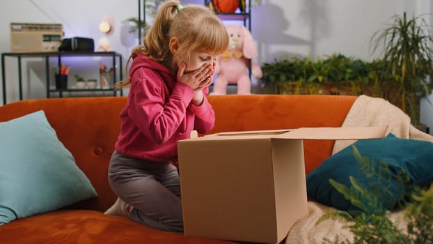 Happy young blonde child kid girl unpacking delivery parcel. Smiling satisfied teen toddler shopper, online shop customer opening cardboard box receiving purchase gift by fast postal shipping at home