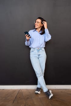 Satisfied hipster girl wear glasses, types text message on mobile phone, enjoys online communication, types feedback, wears denim shirt, isolated on black studio background. Full length