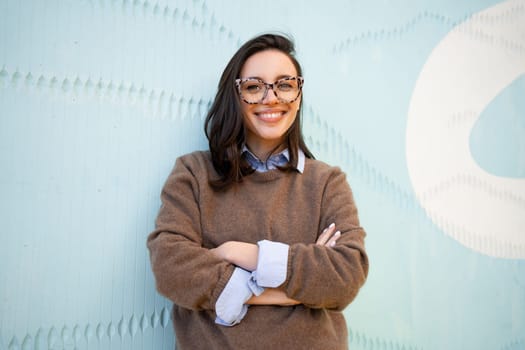 Happy woman in glasses outdoor on blue color background. Positive people concept. Smiling girl in eyeglasses looking at camera hands crossed on chest.