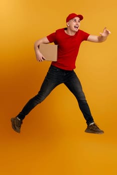 Delivery man in red cap, t-shirt holding cardboard box and jumping isolated on orange background