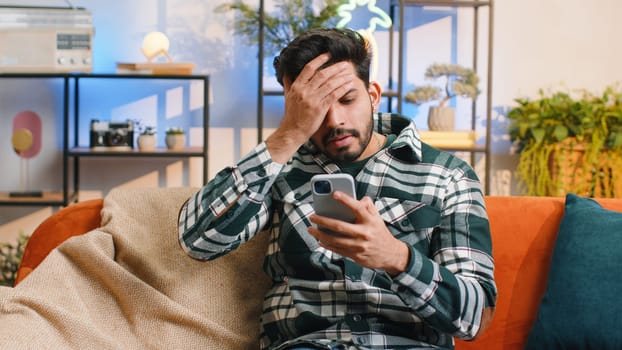 Sad displeased indian man use smartphone typing browsing, loses becoming surprised sudden lottery results, bad news, fortune loss, fail, deadline, virus. Young hindu guy at home living room on couch