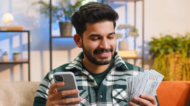 Planning family budget. Smiling indian man counting money cash, use smartphone calculate domestic bills at home room. Joyful hindu guy satisfied of income and saves money for planned vacation, gifts