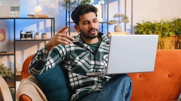 Indian man sitting on couch looking at camera, making video webcam conference call with friends or family, enjoying pleasant conversation. Young hindu guy laughing waving hello at home room