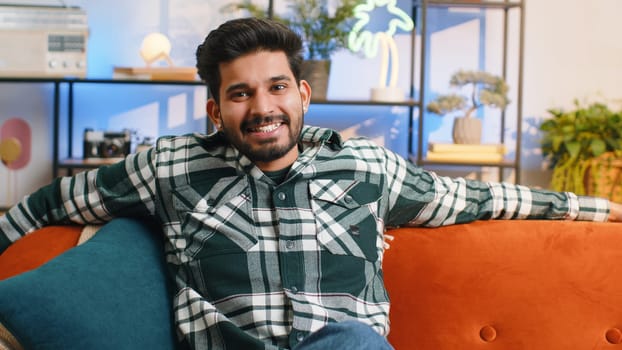 Portrait of happy calm indian man at home couch smiling friendly, glad expression looking away dreaming resting, relaxation feel satisfied concept good news, celebrate win. Hindu guy in living room
