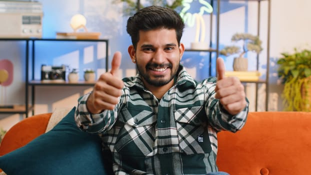 Like. Happy excited indian man looking approvingly at camera showing thumbs up, like sign positive something, good great news, positive feedback. Young hindu guy sitting on couch at home living room