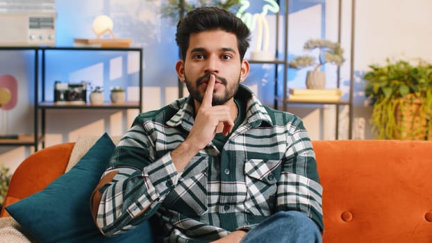 Shh be quiet, hush please. Indian man with presses index finger to lips makes silence hush gesture sign do not tells gossip secret. Young hindu guy at modern home living room sitting on couch. Gossip