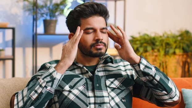 Displeased indian man rubbing temples to cure headache problem, suffering from tension and migraine, stress grimacing in pain high blood pressure at home room apartment. Hindu guy on couch. Lifestyles