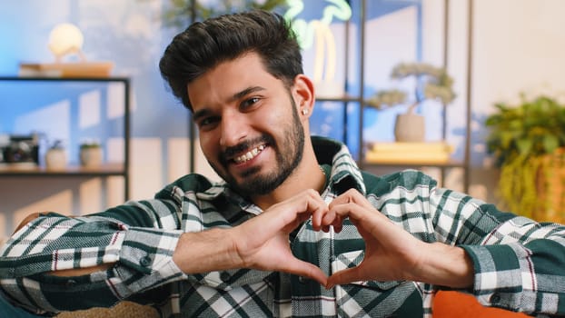 I love you. Happy indian man at home living room couch makes symbol of love, showing heart sign to camera, express romantic feelings express sincere positive feelings. Charity, gratitude, donation