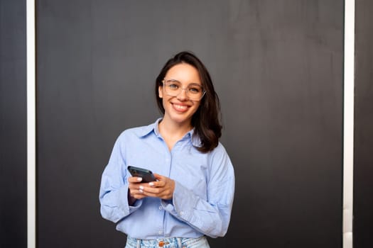 Portrait smiling woman holding smartphone in hand looking at camera, wear glasses, types text message on mobile phone, enjoys online communication, on black studio background. Communication concept