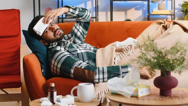 Indian ill man suffering from cold or allergy lying on sofa in living room at home apartment. Sick hindu guy with towel on head blows sneezes wipes snot into napkin. Coronavirus quarantine, pandemic