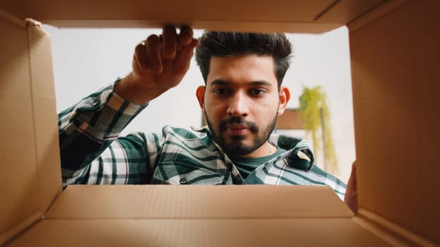 Inside view from box. Indian man unpacking delivery parcel at home. Smiling satisfied hindu guy shopper, online shop customer opening cardboard receiving purchase gift by fast postal shipping order
