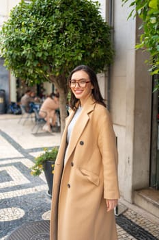 Friendly smiling attractive woman leaving building on background green hedge in the street of European city. Happy confident woman in glasses dressed stylish trench coat smiling looking at camera